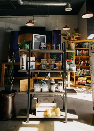 A cozy bookshop corner featuring a display shelf with various items such as coffee beans, snacks, and merchandise. Several potted plants add greenery, while books are neatly arranged on shelves in the background. Warm lighting from hanging lamps overhead gives a comforting ambiance.
