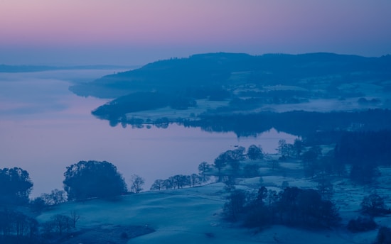 A serene landscape of rolling hills and a wide, calm river or lake reflecting the soft colors of a pastel sky. Mist hovers over the water and trees are scattered across the terrain, contributing to the tranquil atmosphere.