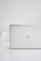 A sleek, modern laptop with a silver finish is placed on a glossy white surface. The iconic logo on the back is visible, indicating the brand. The background is minimalistic, creating a clean and tech-focused atmosphere.