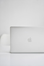 A sleek, modern laptop with a silver finish is placed on a glossy white surface. The iconic logo on the back is visible, indicating the brand. The background is minimalistic, creating a clean and tech-focused atmosphere.