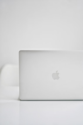 A sleek, modern laptop with a silver finish is placed on a glossy white surface. The iconic logo on the back is visible, indicating the brand. The background is minimalistic, creating a clean and tech-focused atmosphere.