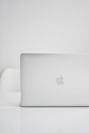 A sleek, modern laptop with a silver finish is placed on a glossy white surface. The iconic logo on the back is visible, indicating the brand. The background is minimalistic, creating a clean and tech-focused atmosphere.