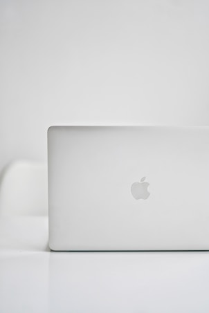 A sleek, modern laptop with a silver finish is placed on a glossy white surface. The iconic logo on the back is visible, indicating the brand. The background is minimalistic, creating a clean and tech-focused atmosphere.