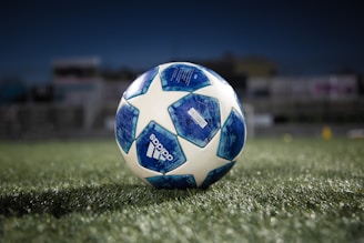 A soccer ball with a blue and white pattern sits on a green grass field under a dark sky, with the brand name visible on the side and a blurred background indicating a stadium or sports field.