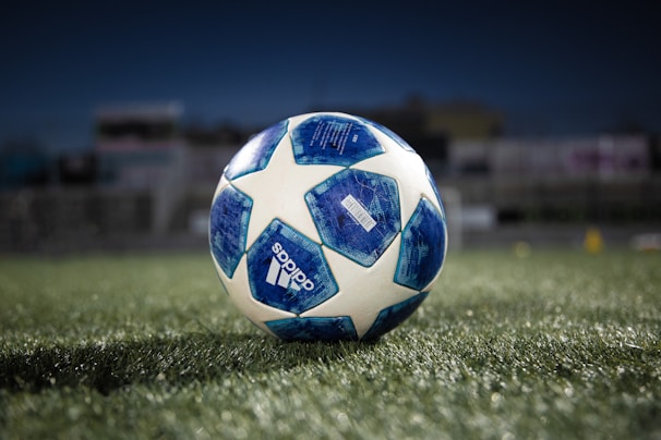 A soccer ball with a blue and white pattern sits on a green grass field under a dark sky, with the brand name visible on the side and a blurred background indicating a stadium or sports field.