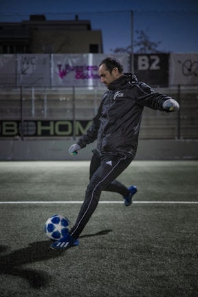 A person wearing a black tracksuit and gloves kicks a blue and white soccer ball on a soccer field. The setting appears to be outdoors with artificial lighting, and there are advertising boards in the background.