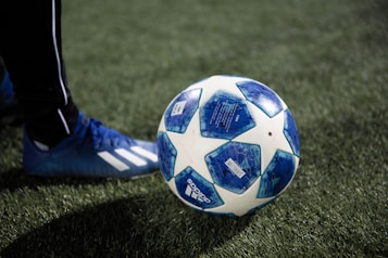 A soccer ball with a blue and white pattern is placed on a green artificial turf field. A person wearing blue soccer shoes with white stripes is positioned nearby, ready to kick the ball.