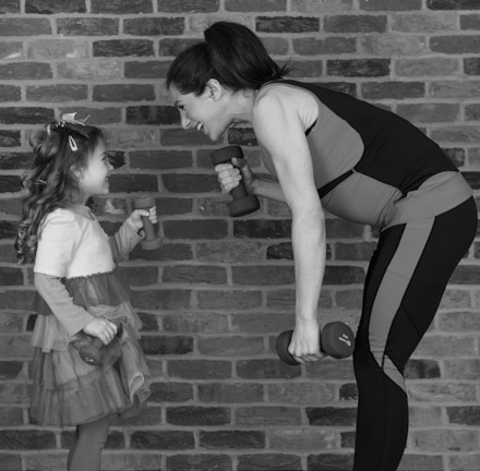 A woman and a young girl face each other, smiling while holding small dumbbells. The woman is leaning forward slightly, wearing athletic clothing, and the child is wearing a dress with layers of ruffles. They appear to be in a playful or exercise setting against a brick wall.