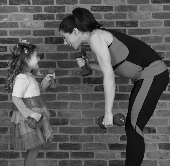 A woman and a young girl face each other, smiling while holding small dumbbells. The woman is leaning forward slightly, wearing athletic clothing, and the child is wearing a dress with layers of ruffles. They appear to be in a playful or exercise setting against a brick wall.
