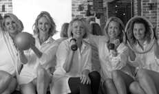 Five women in white shirts sit together holding fitness equipment such as a ball, dumbbells, and exercise rings. They are positioned against a backdrop of brick walls and mirrors, creating a lively and cheerful atmosphere.