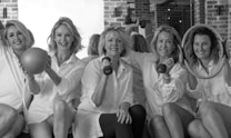 Five women in white shirts sit together holding fitness equipment such as a ball, dumbbells, and exercise rings. They are positioned against a backdrop of brick walls and mirrors, creating a lively and cheerful atmosphere.