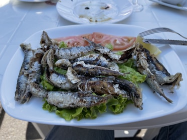 A plate of grilled fish, likely sardines, is served on a bed of green lettuce with slices of fresh tomato and a lemon wedge. The fish have crispy, charred skins and appear to be freshly cooked. The plate rests on a white table, and there are some empty dishes in the background, suggesting a meal setting.