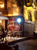 A dimly lit street scene features a cafe with outdoor seating where a few people are sitting at tables. A striking mural of a face with tribal-style white markings dominates the scene. The building next to the cafe has ornate, arched windows with wrought iron details, and there's a motorcycle parked nearby.