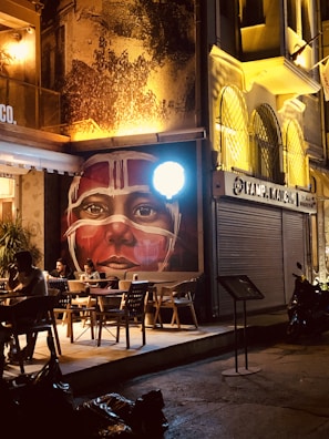 A dimly lit street scene features a cafe with outdoor seating where a few people are sitting at tables. A striking mural of a face with tribal-style white markings dominates the scene. The building next to the cafe has ornate, arched windows with wrought iron details, and there's a motorcycle parked nearby.