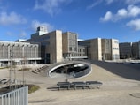 A modern architectural building with geometric shapes and large glass windows. The structure is surrounded by an open plaza with benches, young trees, and a unique ramp structure leading underground. The sky is clear with a few clouds, adding to the tranquility of the environment.