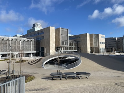 A modern architectural building with geometric shapes and large glass windows. The structure is surrounded by an open plaza with benches, young trees, and a unique ramp structure leading underground. The sky is clear with a few clouds, adding to the tranquility of the environment.