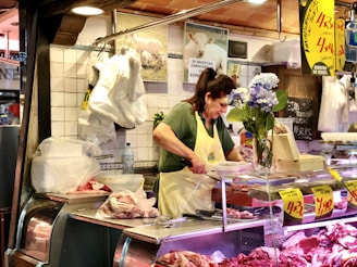 A woman is working at a butcher shop counter, surrounded by various cuts of meat displayed on the glass counter. She is wearing a yellow apron and seems focused on her task. Behind her, there are signs and advertisements for the meat, as well as hanging bags and a wall decorated with tiles. A bouquet of blue flowers adds a touch of color to the scene.