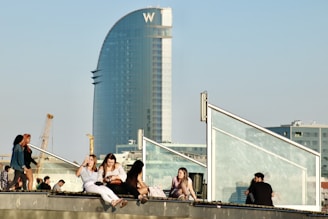 A group of people is sitting and standing on a rooftop with clear glass barriers. In the background, a large, modern, sail-shaped skyscraper stands against a clear blue sky. Construction cranes are visible, adding an urban and developmental touch.
