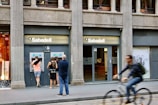 A street scene featuring a bank called Sabadell with two ATM machines and several people interacting with them. The building has a classic architectural style with large columns. A person on a bicycle is riding past in the foreground.