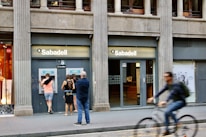 A street scene featuring a bank called Sabadell with two ATM machines and several people interacting with them. The building has a classic architectural style with large columns. A person on a bicycle is riding past in the foreground.