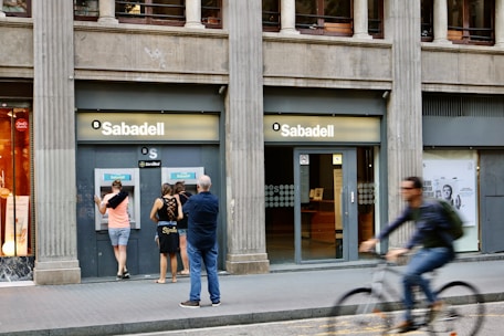 A street scene featuring a bank called Sabadell with two ATM machines and several people interacting with them. The building has a classic architectural style with large columns. A person on a bicycle is riding past in the foreground.