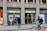 A street scene featuring a bank called Sabadell with two ATM machines and several people interacting with them. The building has a classic architectural style with large columns. A person on a bicycle is riding past in the foreground.