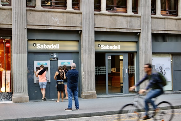 A street scene featuring a bank called Sabadell with two ATM machines and several people interacting with them. The building has a classic architectural style with large columns. A person on a bicycle is riding past in the foreground.