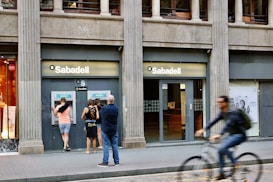 A street scene featuring a bank called Sabadell with two ATM machines and several people interacting with them. The building has a classic architectural style with large columns. A person on a bicycle is riding past in the foreground.
