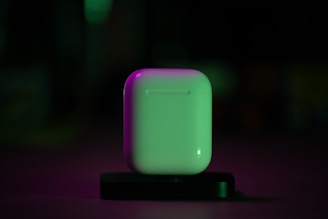 A white charging case, resembling one used for wireless earbuds, is illuminated with green and purple lighting, creating a dramatic and stylish effect. It is centered on a dark background, enhancing the visibility of the object.