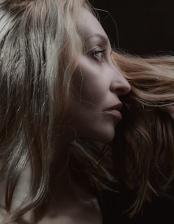 A profile view of a person with long, light-colored hair. The focus is on the contours of the face and the texture of the hair, set against a dark background.