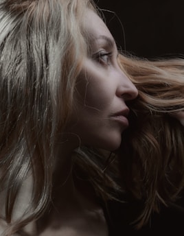 A profile view of a person with long, light-colored hair. The focus is on the contours of the face and the texture of the hair, set against a dark background.