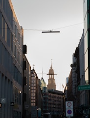A city street view with tall buildings on either side leading toward a distant clock tower bathed in soft sunlight. One building prominently displays a sign reading 'ALSTERHAUS'. Traffic signs and a hotel sign are visible in the foreground, and the street is relatively calm.
