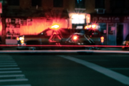 A street scene at night featuring a blurred emergency response vehicle with flashing red lights. The background includes indistinct buildings and storefronts, illuminated by the glow of the vehicle