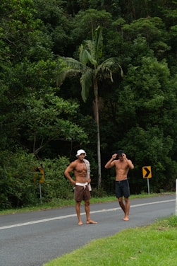 Two shirtless men stand on the side of a paved road, surrounded by dense green foliage and tall trees. Both men wearing hats, with one adjusting his binoculars. The scene appears to be outdoors in a tropical or forested area. To their left are yellow traffic signs with black arrows, indicating a curve in the road.