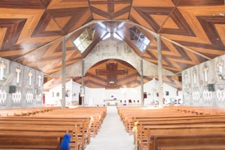A large church interior with wooden pews arranged in rows. The ceiling features intricate wooden designs forming geometric patterns. The walls display multiple crosses and modern light fixtures. The altar is centrally located at the far end, partially visible in the bright interior.