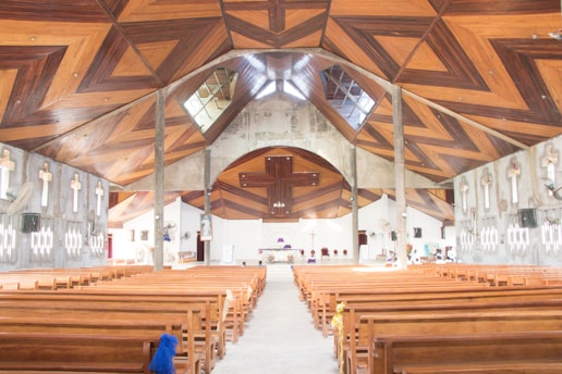 A large church interior with wooden pews arranged in rows. The ceiling features intricate wooden designs forming geometric patterns. The walls display multiple crosses and modern light fixtures. The altar is centrally located at the far end, partially visible in the bright interior.