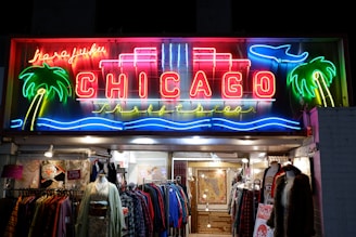A vibrant neon sign depicting the word 'CHICAGO' is lit up against a dark background. The sign is adorned with colorful elements such as palm trees, a wave pattern, and a plane. Below the sign, a shop entrance is visible with racks of colorful clothing and mannequins. The shop appears inviting and showcases a mix of garments.