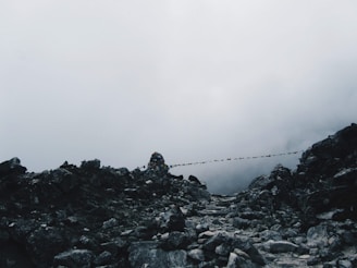 A rugged mountain trail covered with rocks and stones, leading into the foggy distance. Small colorful prayer flags are strung across the rocky path, adding a touch of color against the grayscale backdrop. The sky is overcast, creating a moody, atmospheric scene.