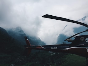 A helicopter is positioned against a backdrop of mist-covered mountains. The scene is dominated by dense clouds that shroud the peaks, creating a dramatic and mysterious atmosphere. The helicopter appears stationary, situated in a lush, green valley.