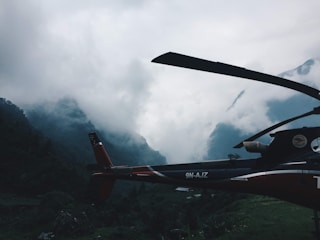 A helicopter is positioned against a backdrop of mist-covered mountains. The scene is dominated by dense clouds that shroud the peaks, creating a dramatic and mysterious atmosphere. The helicopter appears stationary, situated in a lush, green valley.
