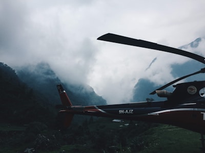 A helicopter is positioned against a backdrop of mist-covered mountains. The scene is dominated by dense clouds that shroud the peaks, creating a dramatic and mysterious atmosphere. The helicopter appears stationary, situated in a lush, green valley.