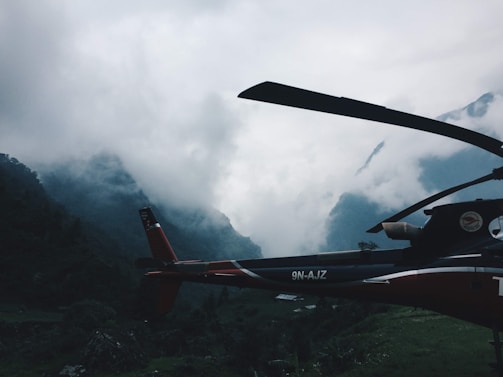 A helicopter is positioned against a backdrop of mist-covered mountains. The scene is dominated by dense clouds that shroud the peaks, creating a dramatic and mysterious atmosphere. The helicopter appears stationary, situated in a lush, green valley.