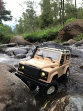 A toy off-road vehicle is situated in a shallow stream surrounded by rocks. The vehicle has a beige color with its headlights turned on, appearing as though it is navigating through a natural setting with trees and greenery in the background.