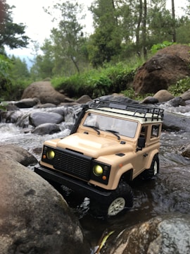 A toy off-road vehicle is situated in a shallow stream surrounded by rocks. The vehicle has a beige color with its headlights turned on, appearing as though it is navigating through a natural setting with trees and greenery in the background.