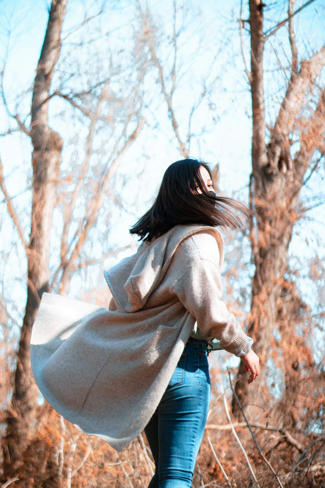 woman in brown long sleeve shirt and blue denim jeans standing near brown trees during daytime