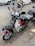 Two motorcycles are parked on the side of a street. The nearest one has a red and white body with chrome details and a wire basket on the front. The other motorcycle is partially visible and seems to have a helmet placed on the seat. Cars and other vehicles are on the road in the background, indicating a busy street environment.