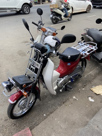 Two motorcycles are parked on the side of a street. The nearest one has a red and white body with chrome details and a wire basket on the front. The other motorcycle is partially visible and seems to have a helmet placed on the seat. Cars and other vehicles are on the road in the background, indicating a busy street environment.