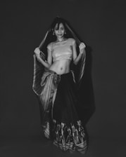 A stylish woman wearing a modernized traditional outfit in black and white, standing confidently against a minimalist dark background.