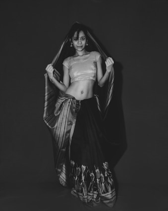 A stylish woman wearing a modernized traditional outfit in black and white, standing confidently against a minimalist dark background.