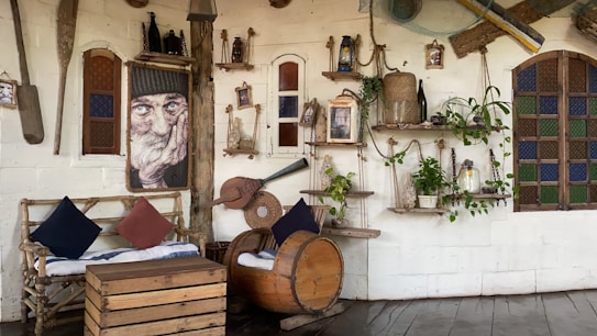 A rustic indoor space featuring a wall adorned with various vintage and nautical-themed decor, including old oars, ropes, bottles, and framed art. A cozy seating area with a wooden couch, adorned with cushions, is complemented by a unique chair made from a barrel. Shelves display plants and lanterns, creating a homely and artistic atmosphere.